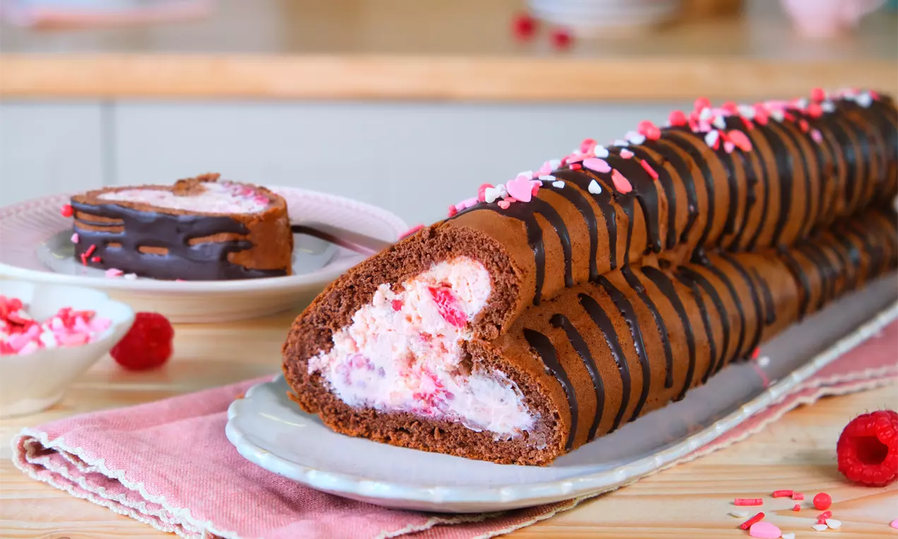 Schokoladige Biskuitrolle in Herzform, gefüllt mit rosa-weißem Himbeer-Sahne-Mix, dekoriert mit Zuckerdekor auf einem langen Teller