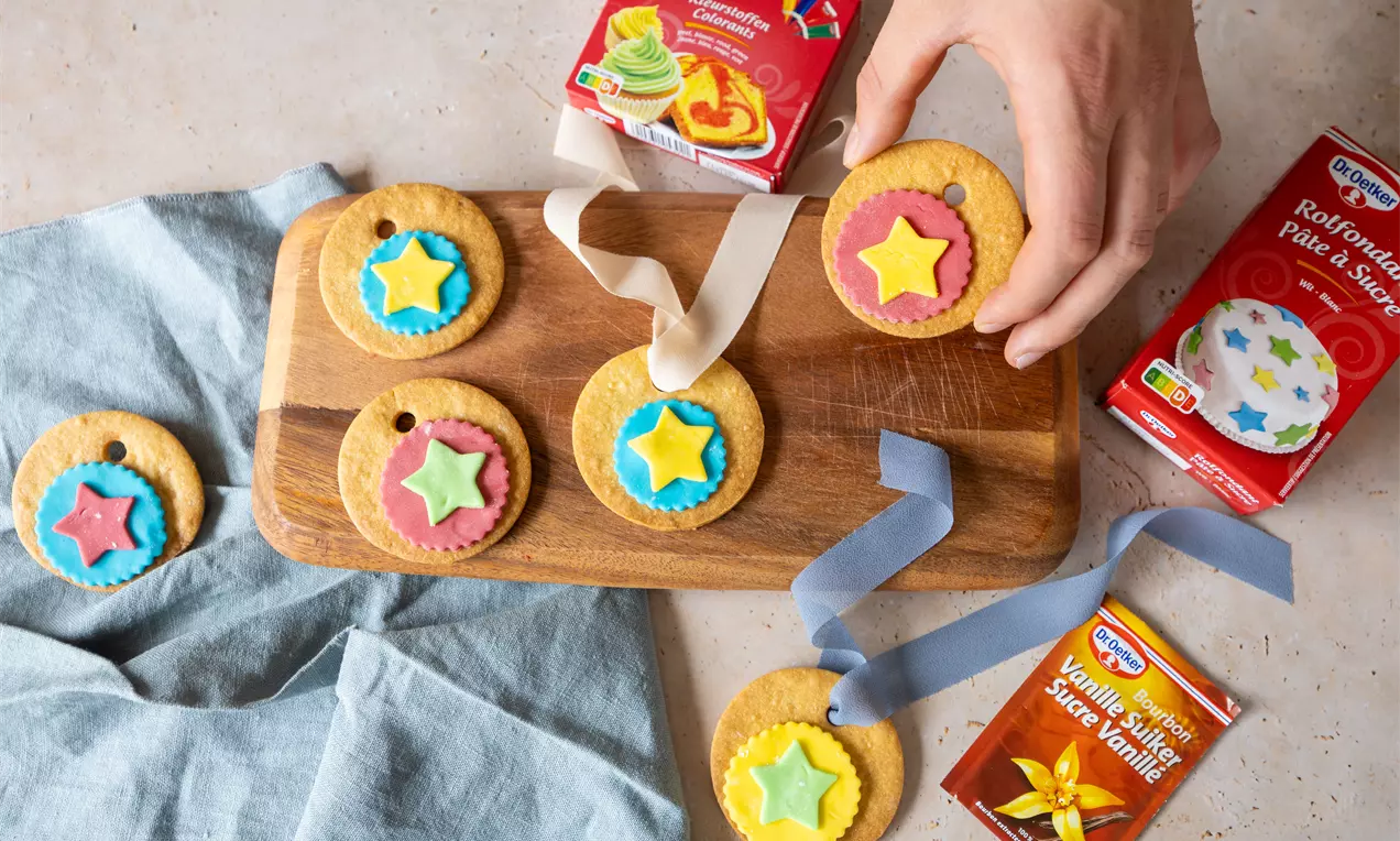 Goudkleurige ronde koekjes met kleurrijke suikersterren en linten op een houten plank, omringd door ingrediënten en een blauwe doek
