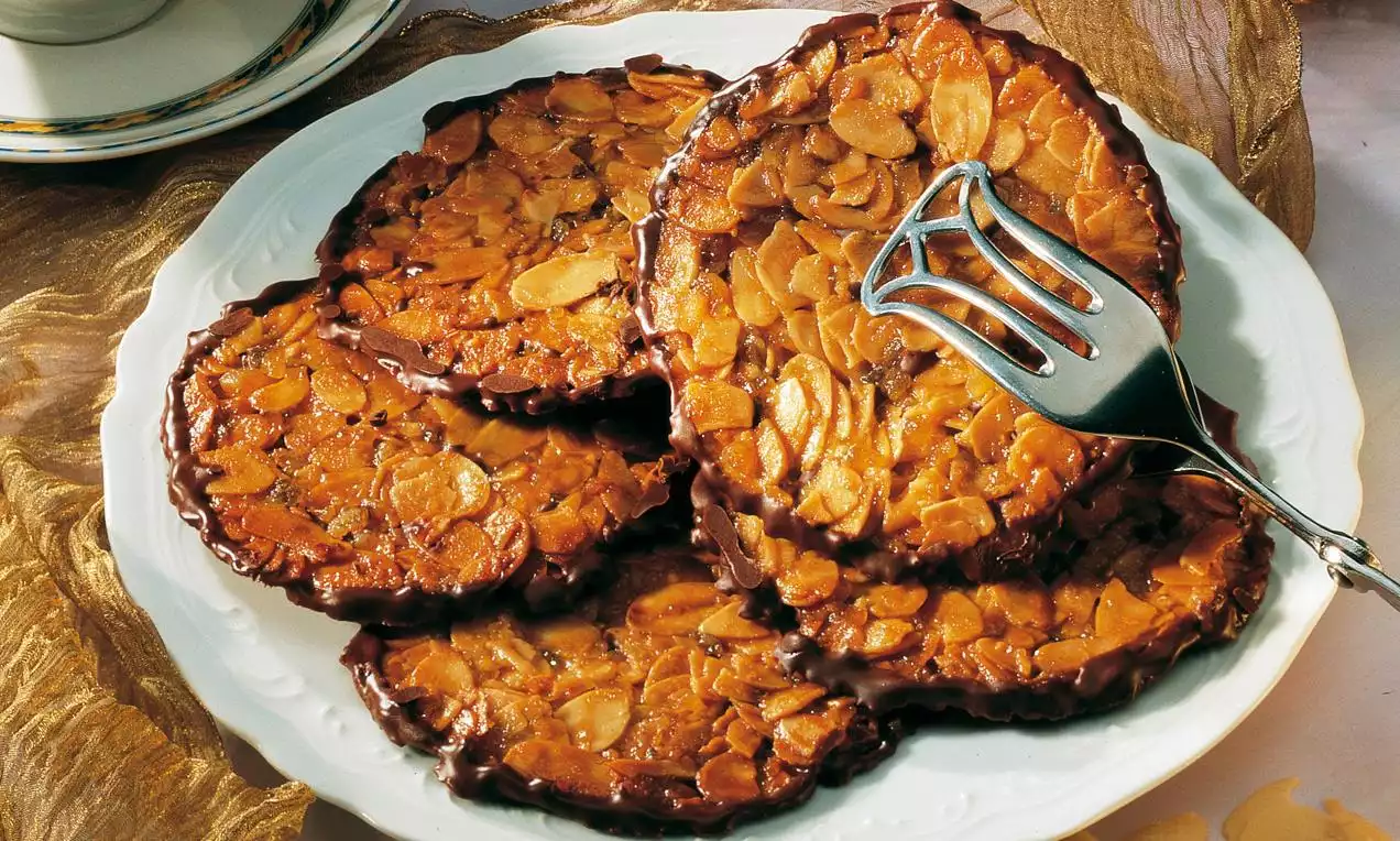 Amandelkoekjes met glanzende karamelkleurige plakjes amandel en een chocoladerand, geordend op een wit bord met een metalen taartschep