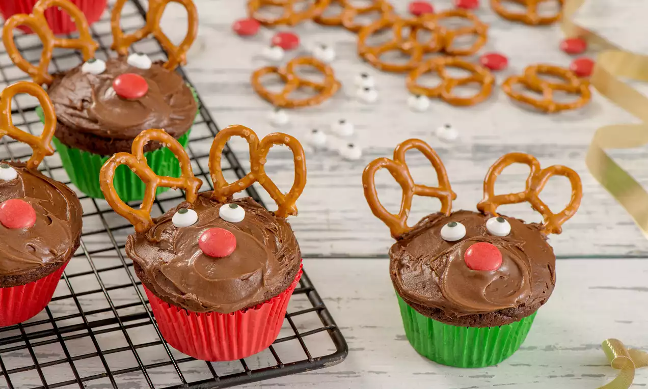 Cupcakes au chocolat glacés, garnis de bretzels dorés en forme de bois, yeux en sucre blancs et nez rouge sur un fond clair