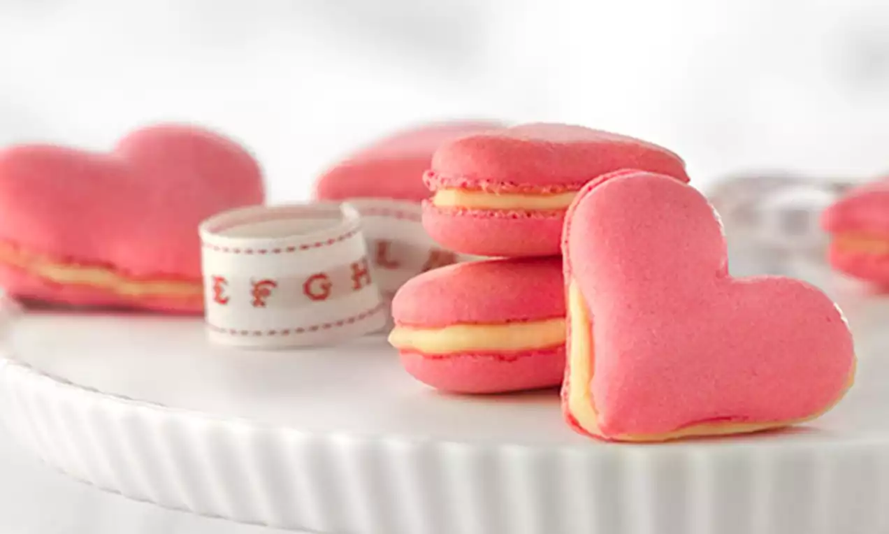 Macarons en forma de corazón de color rojo suave, con relleno cremoso amarillo claro, sobre un plato blanco y cinta decorativa