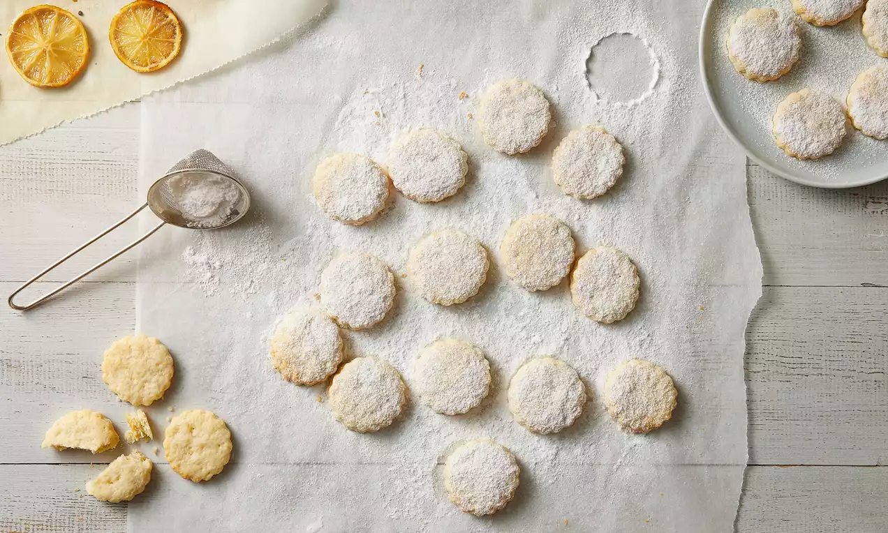 Yuvarlak, altın rengi limonlu kurabiyeler beyaz pudra şekeriyle kaplı, pişirme kağıdında düzenli şekilde sıralanmış ve yanlarında çanakta birkaç parça kurabiye