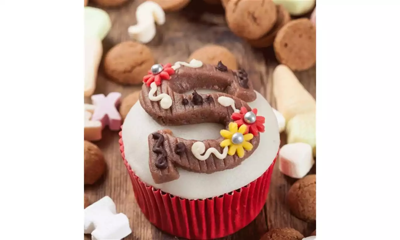 Witte cupcake met rode papieren houder, bovenop een bruine chocoladeletter versierd met bloemen van glazuur en suikerparels, omringd door strooigoed