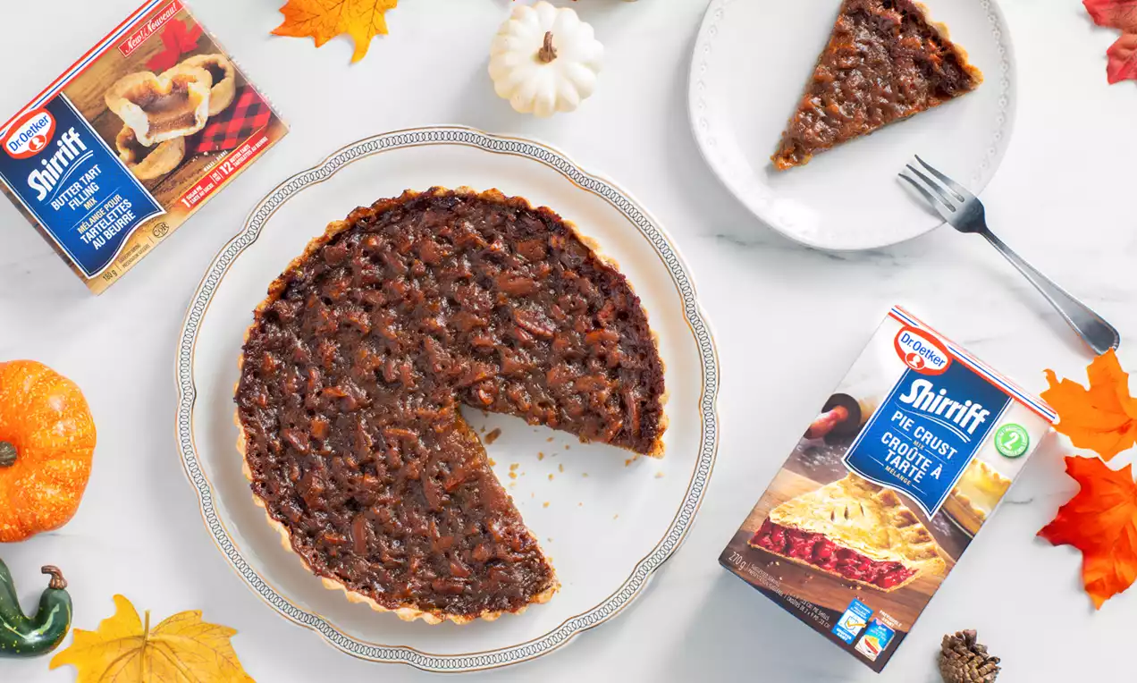 Pumpkin pecan and butter tart pie with a glossy caramelized top on a white plate, surrounded by autumn leaves and miniature pumpkins
