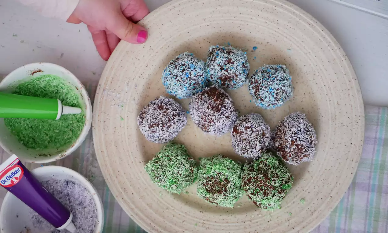Chokladbollar täckta av kokos färgad i grönt, blått och vitt arrangerade på en beige tallrik, omgivna av skålar med färgat kokos och en grön dekorationspenna