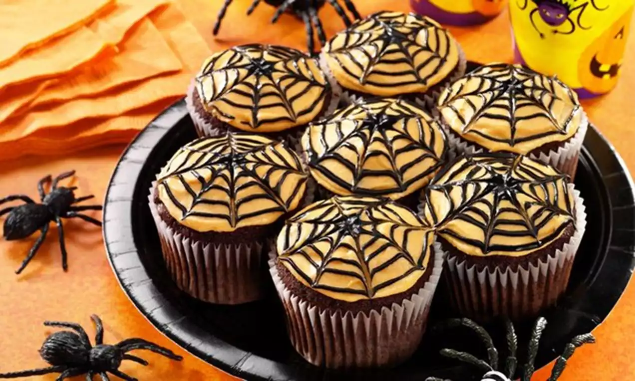 Čokoladni cupcakes prekriveni kremom boje karamele ukrašeni paukovom mrežom od tamne glazure na crnom tanjiru s Halloween dekoracijom