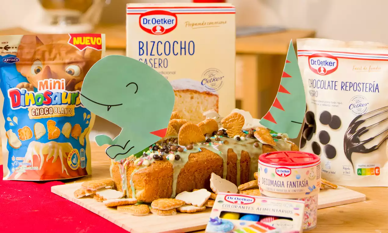 Bizcocho rectangular dorado con glaseado blanco, decorado con galletas, chispas de chocolate y detalles de dinosaurio en papel