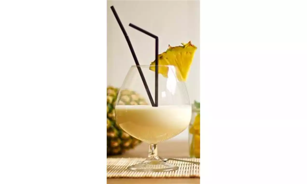 Schuimige lichtgele ananasshake in een rond glas met twee donkere rietjes en een verse ananasschijf op de rand van het glas