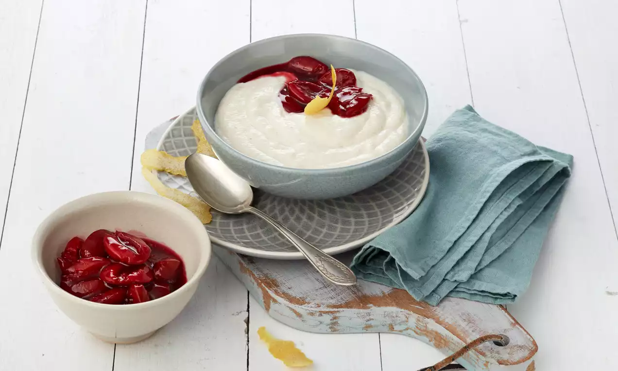 Cremiger, weißer Grießbrei in einer graublauen Schale, garniert mit roten Kompottfrüchten und Zitronenzesten auf grauem Teller mit Stoffserviette