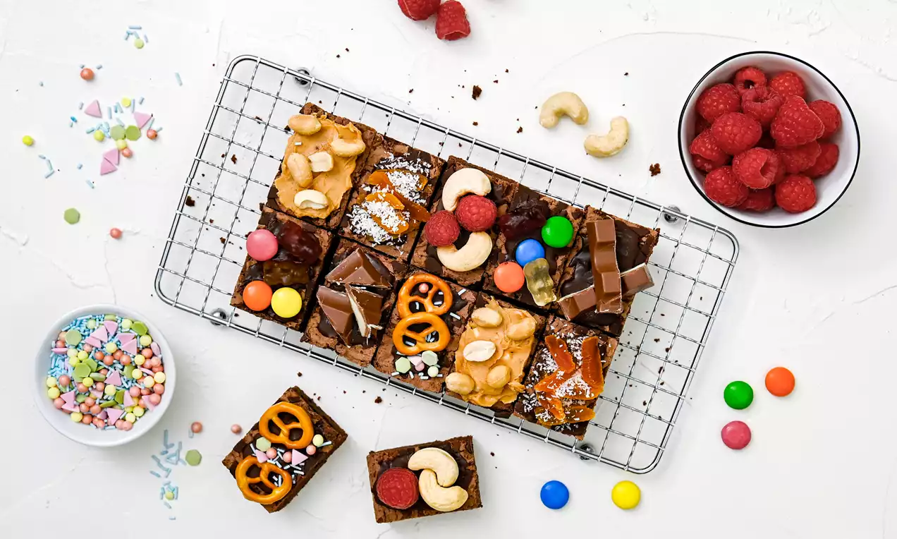 Brownie-Happen auf silbernem Gitterrost, verziert mit bunten Süßigkeiten, Pretzeln, Cashews, Himbeeren und Schokolade