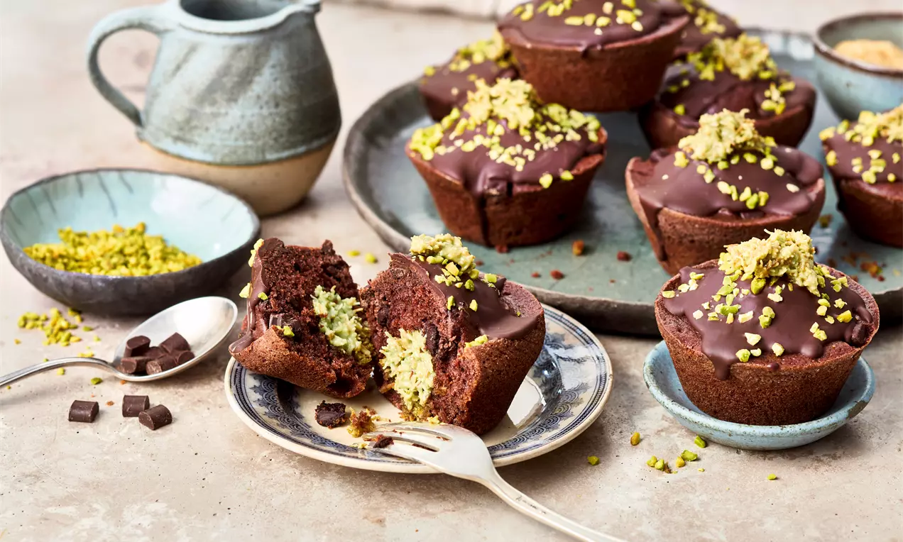 Rich chocolate muffins topped with glossy dark chocolate glaze and crushed pistachios, arranged on ceramic plates with scattered toppings