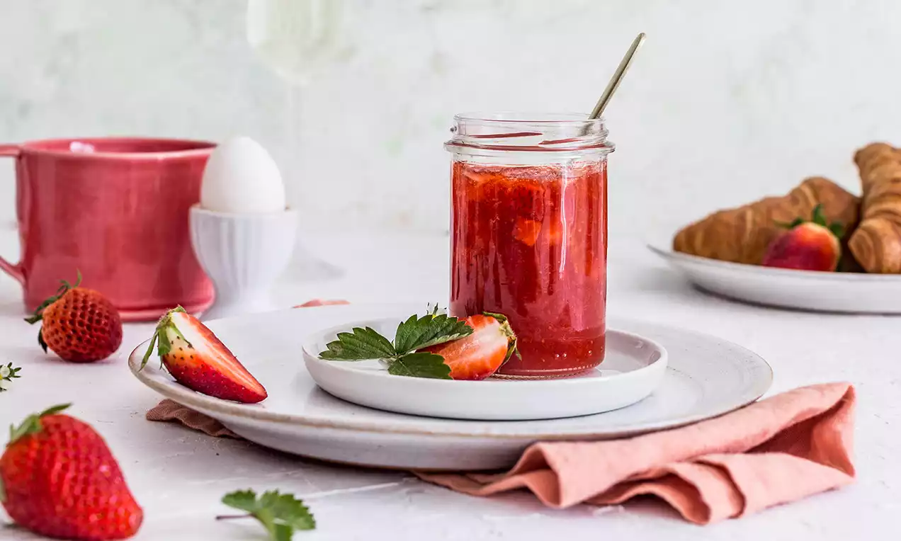 Sklenice se sytě červeným jahodovým džemem s proseccem, usazená na bílém talíři, obklopená čerstvými jahodami, růžovou látkou a pečivem