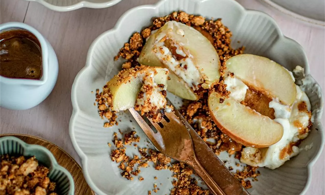 Златисто-жълти половинки печени ябълки с кремообразен топинг, разположени върху хрупкав кафяв крамбъл в светла чиния с десертна вилица