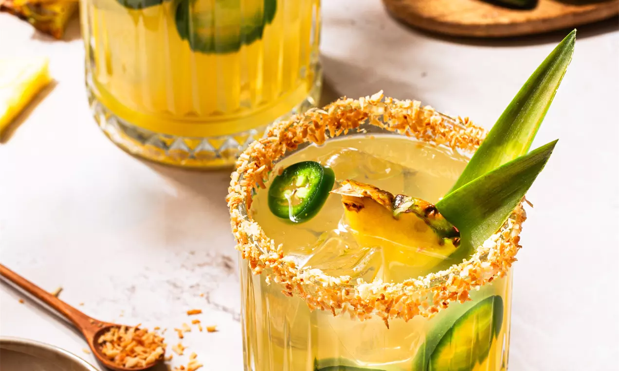 Picture - Two golden-yellow margaritas in textured glass tumblers, rimmed with toasted coconut, garnished with jalapeño slices and pineapple leaves