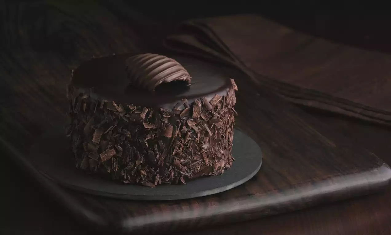 Mini dark chocolate cake with glossy smooth ganache top, curled chocolate garnish, and sides coated in flaky chocolate shards on a slate plate