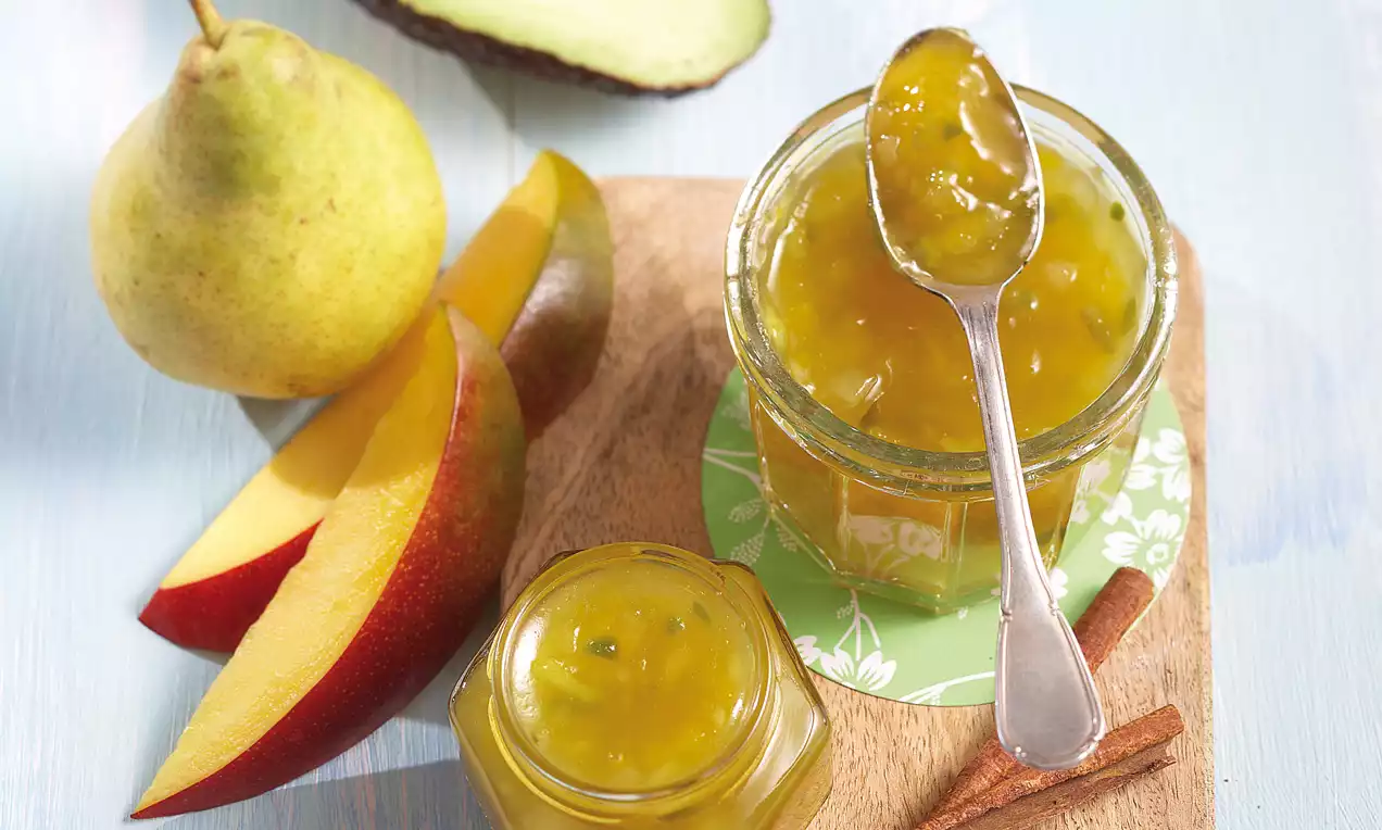 Confiture dorée et brillante aux morceaux de mangue, poire, et avocat, dans des pots en verre sur une planche avec bâtons de cannelle