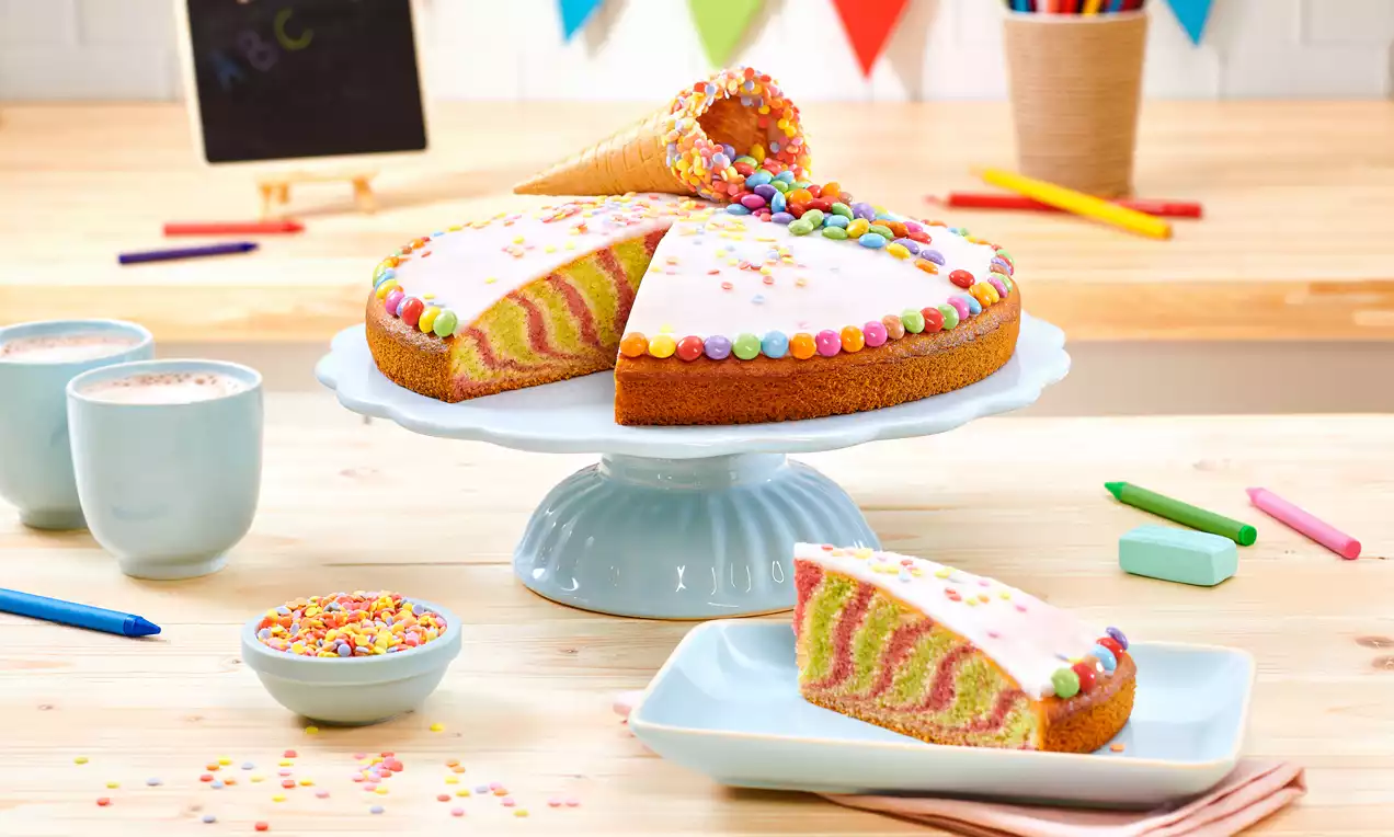 Bunter Kuchen mit rosa-grünem Zebramuster, weißer Glasur und runder Dekoration, auf einem hellblauen Kuchenteller mit Schultüte
