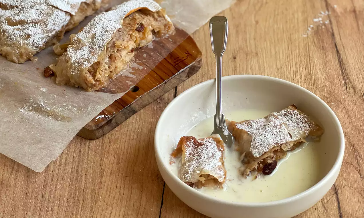 Zlatnosmeđi Apfelstrudel punjen voćem, posut šećerom u prahu, narezan na drvenoj dasci uz tanjir s vanilin kremom