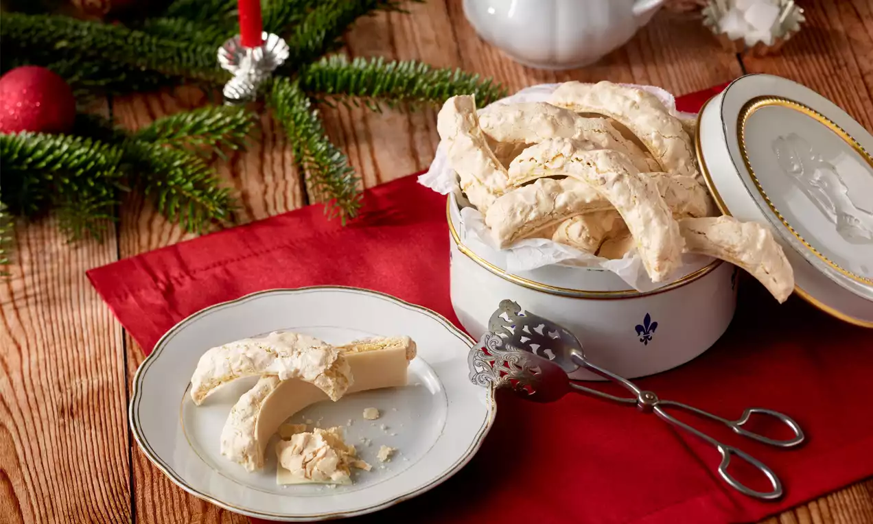 Helle, leicht gebogene Mandelbögen mit feiner, brüchiger Textur auf einem Teller und in einer weißen Dose, umgeben von Tannenzweigen