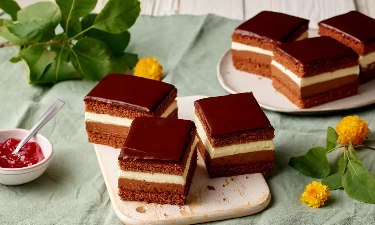 Rechteckige Schokomousse-Schnitten mit glänzender Schokoladenglasur, heller Cremefüllung und dunklem Boden auf Holzbrett mit Blumen dekoriert