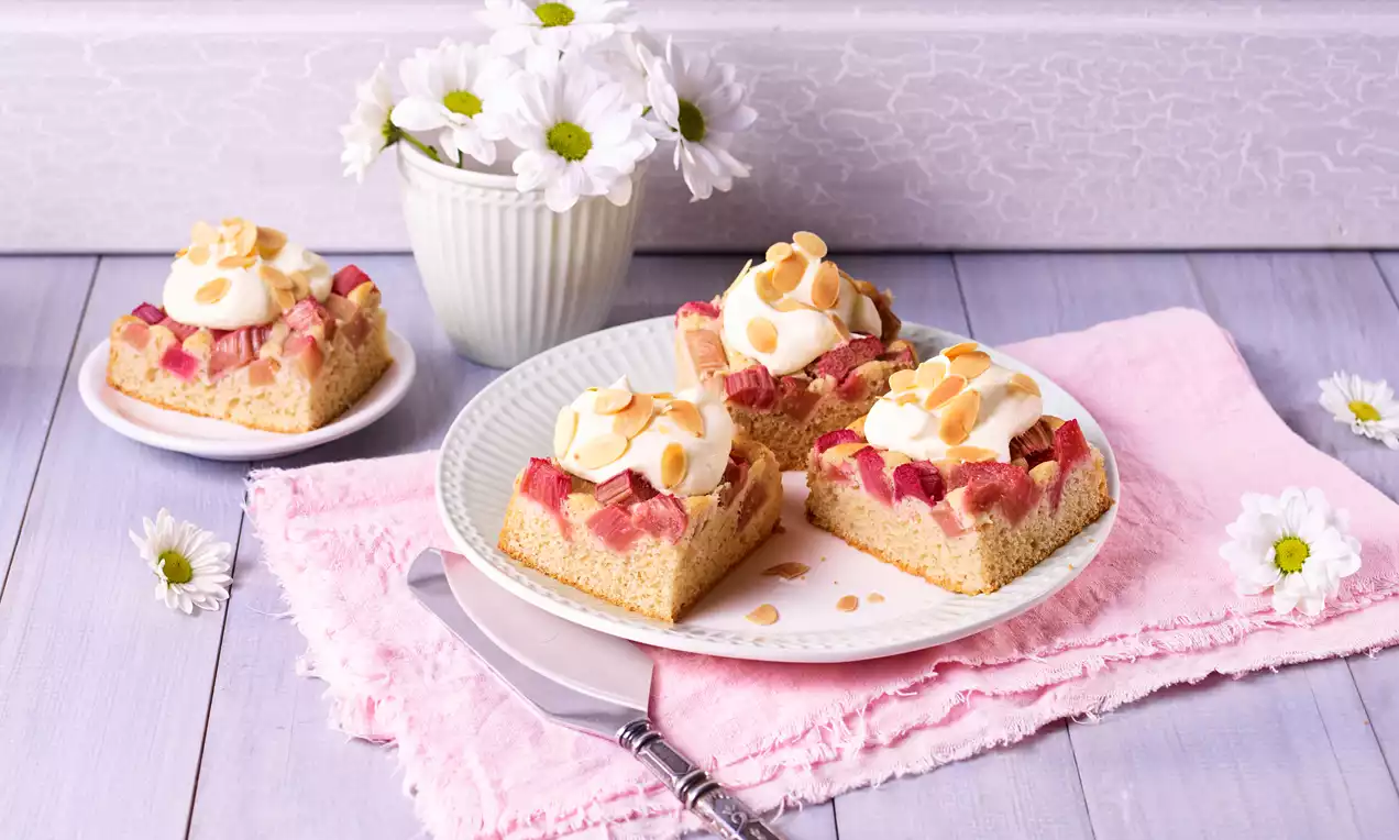 Carrés dorés moelleux garnis de morceaux de rhubarbe rose, crème blanche onctueuse et éclats d’amandes sur une assiette blanche