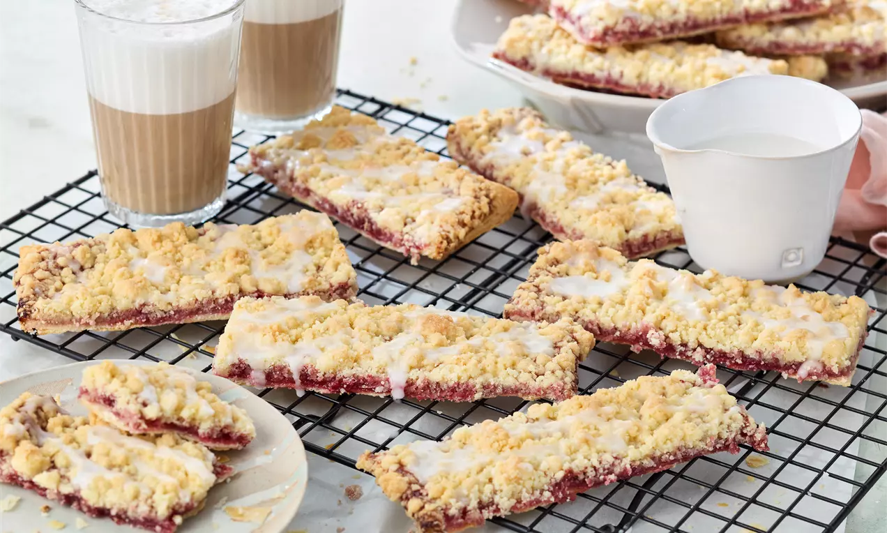 Prasselkuchen mit goldgelber Streuselkruste und hellweißem Zuckerguss auf dunkler Drahtablage, daneben Milchkaffee und ein Teller