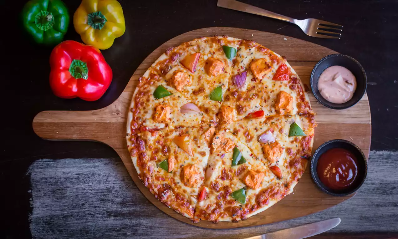Round tandoori pizza with golden crust, melted cheese, orange chicken pieces, green and red bell peppers on a wooden board with sauces