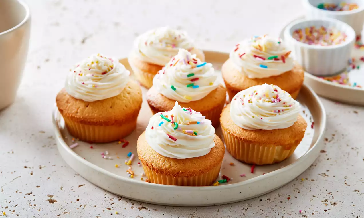 Cupcakes dorés à la vanille avec une crème blanche onctueuse en spirale, parsemés de vermicelles colorés sur une assiette beige