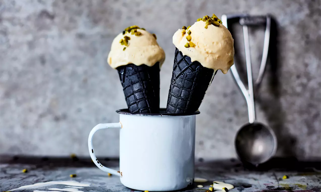 Домашен ванилов сладолед с мека кремообразна текстура в черни фунийки, поръсен с натрошени шамфъстъци, подреден в бяла метална чаша