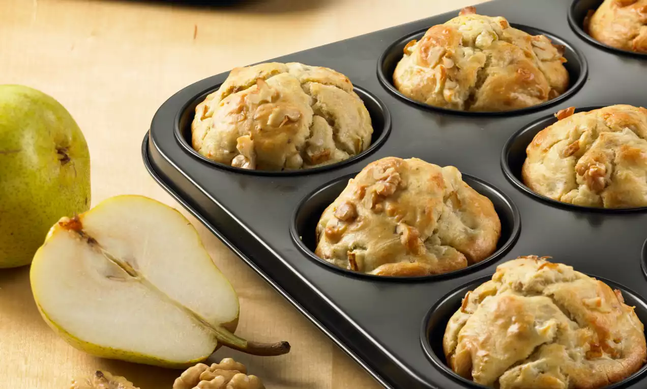 Muffins dorés aux contours irréguliers avec des morceaux de roquefort et de poire dans un moule noir sur une table avec des noix et une poire ouverte