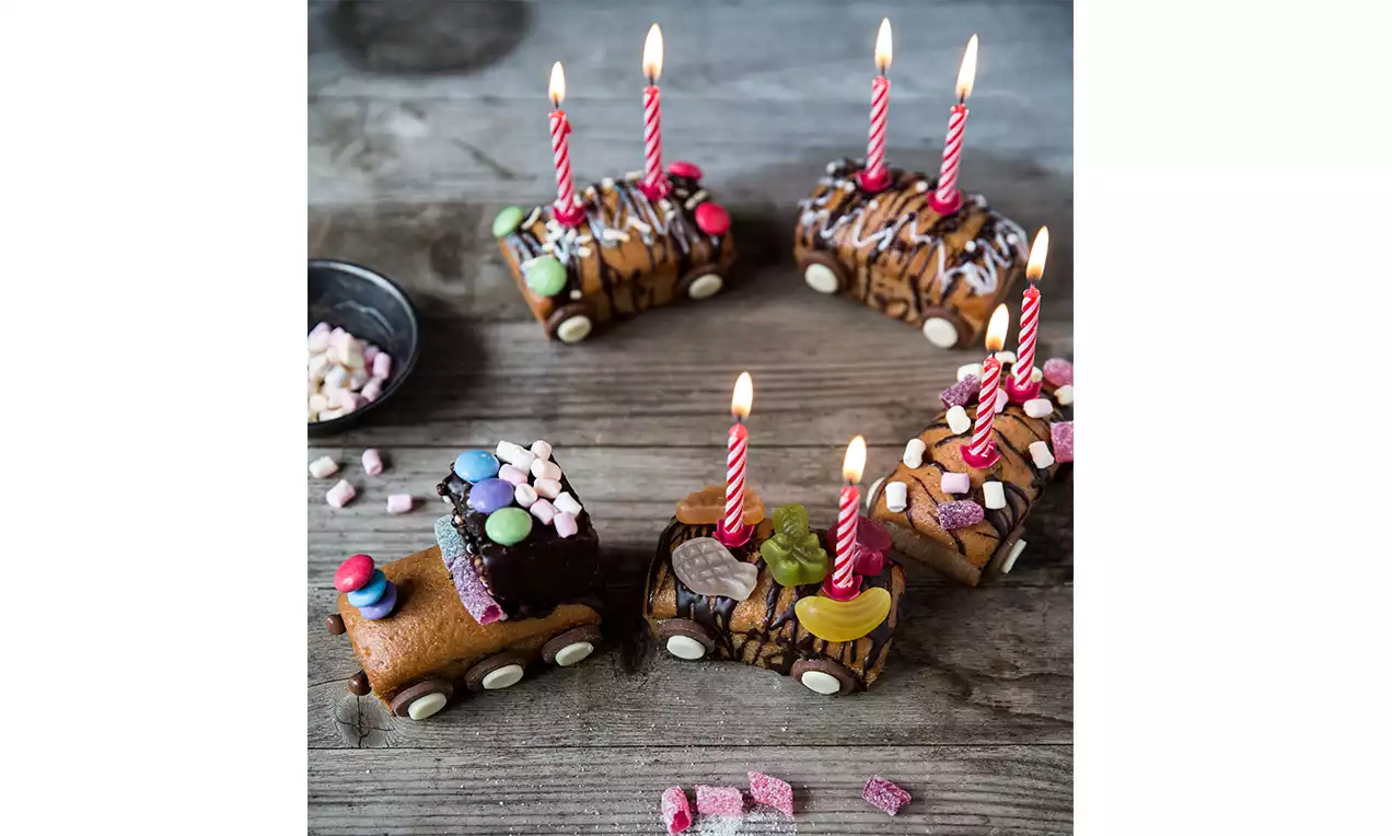 Trenino composto da dolci rettangolari al cioccolato decorati con candele rosse, ruote di zucchero bianco, caramelle colorate e zucchero glassato