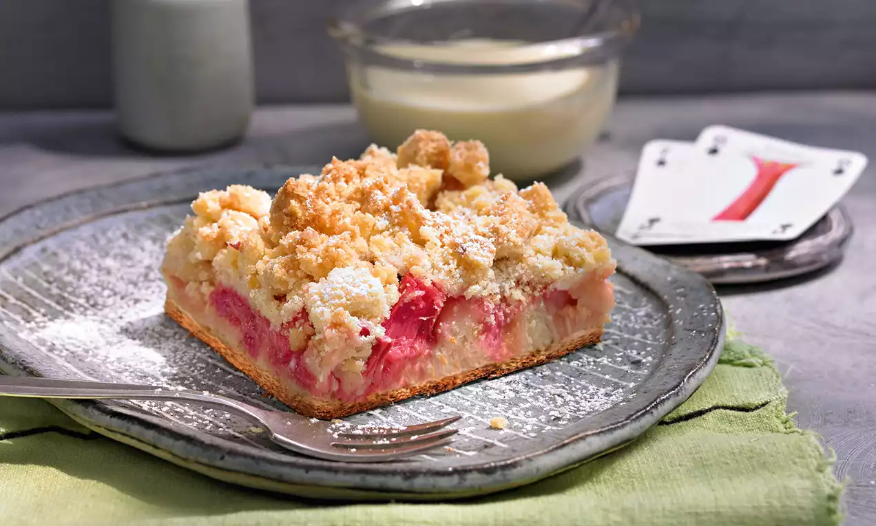 Ein rechteckiges Stück Kuchen mit goldbraunen Streuseln, rosarotem Rhabarber-Erdbeer-Belag, auf silbrigem Teller mit Puderzucker bestäubt