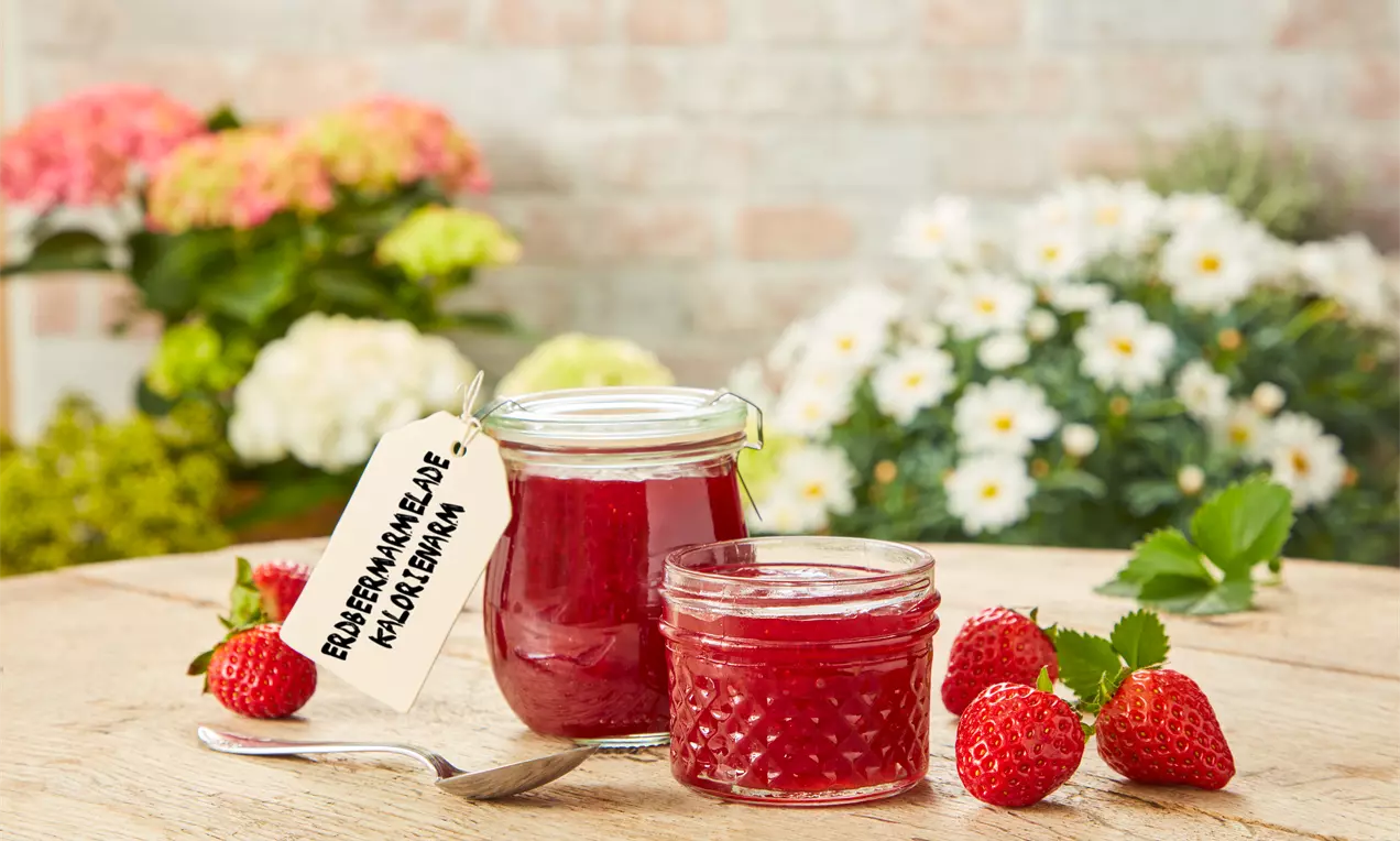 Glas mit kräftig roter Erdbeermarmelade und handbeschriebenem Etikett auf Holzuntergrund mit frischen Erdbeeren und Gartenblumen im Hintergrund