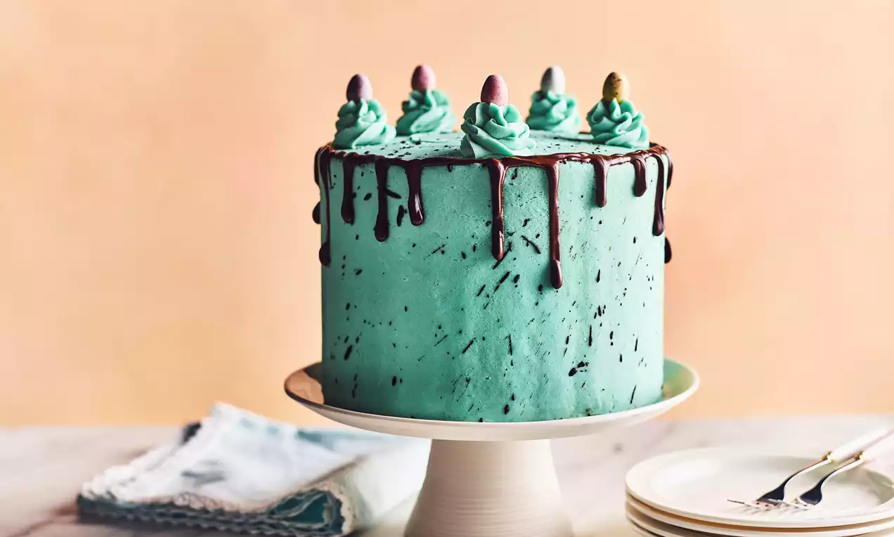 A tall, round speckled cake with smooth turquoise frosting, chocolate drips, teal swirls, and pastel mini eggs on a white cake stand
