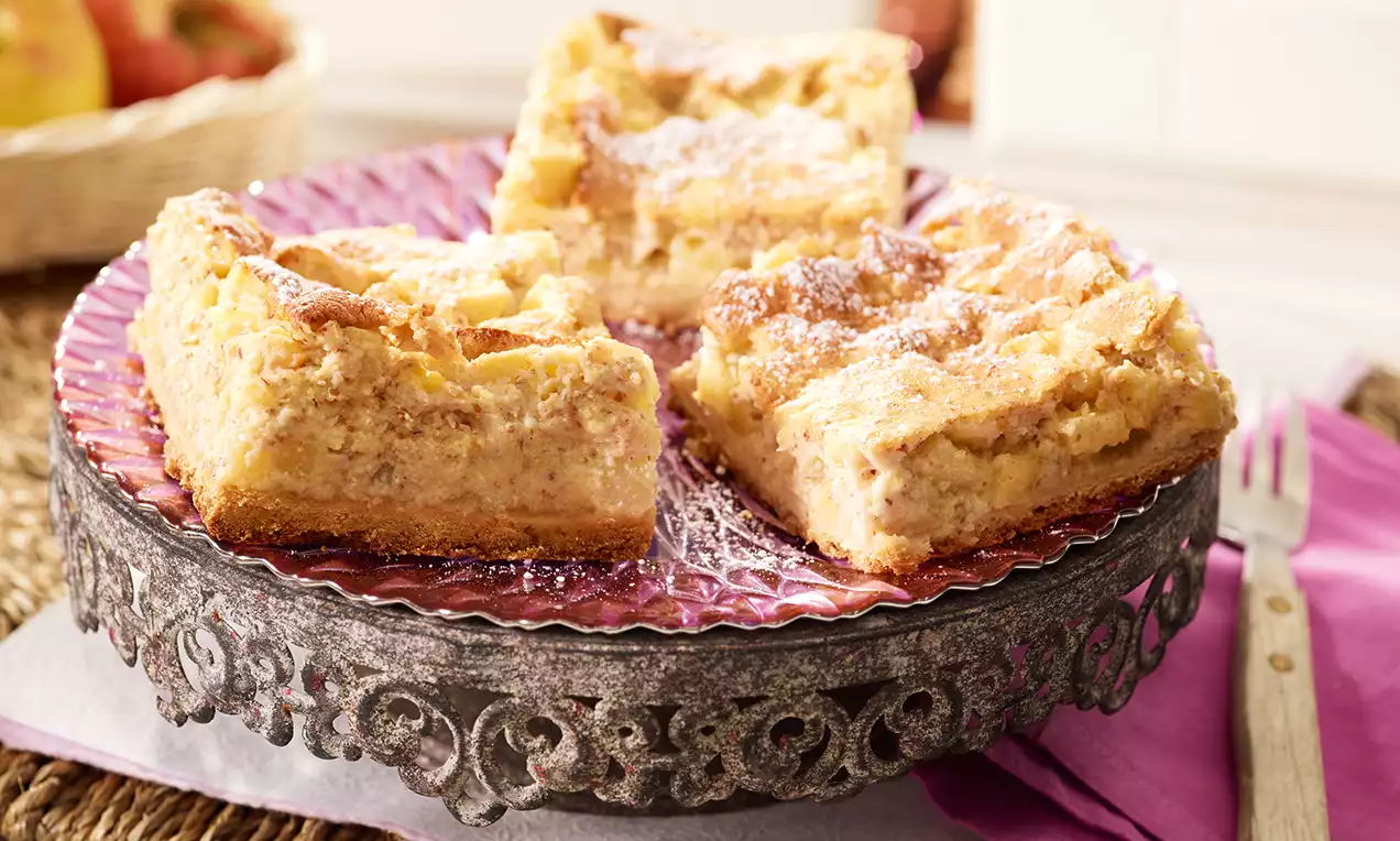 Carrés dorés de gâteau soufflé aux pommes avec une texture moelleuse, saupoudrés de sucre glace sur un plateau rose à bord orné