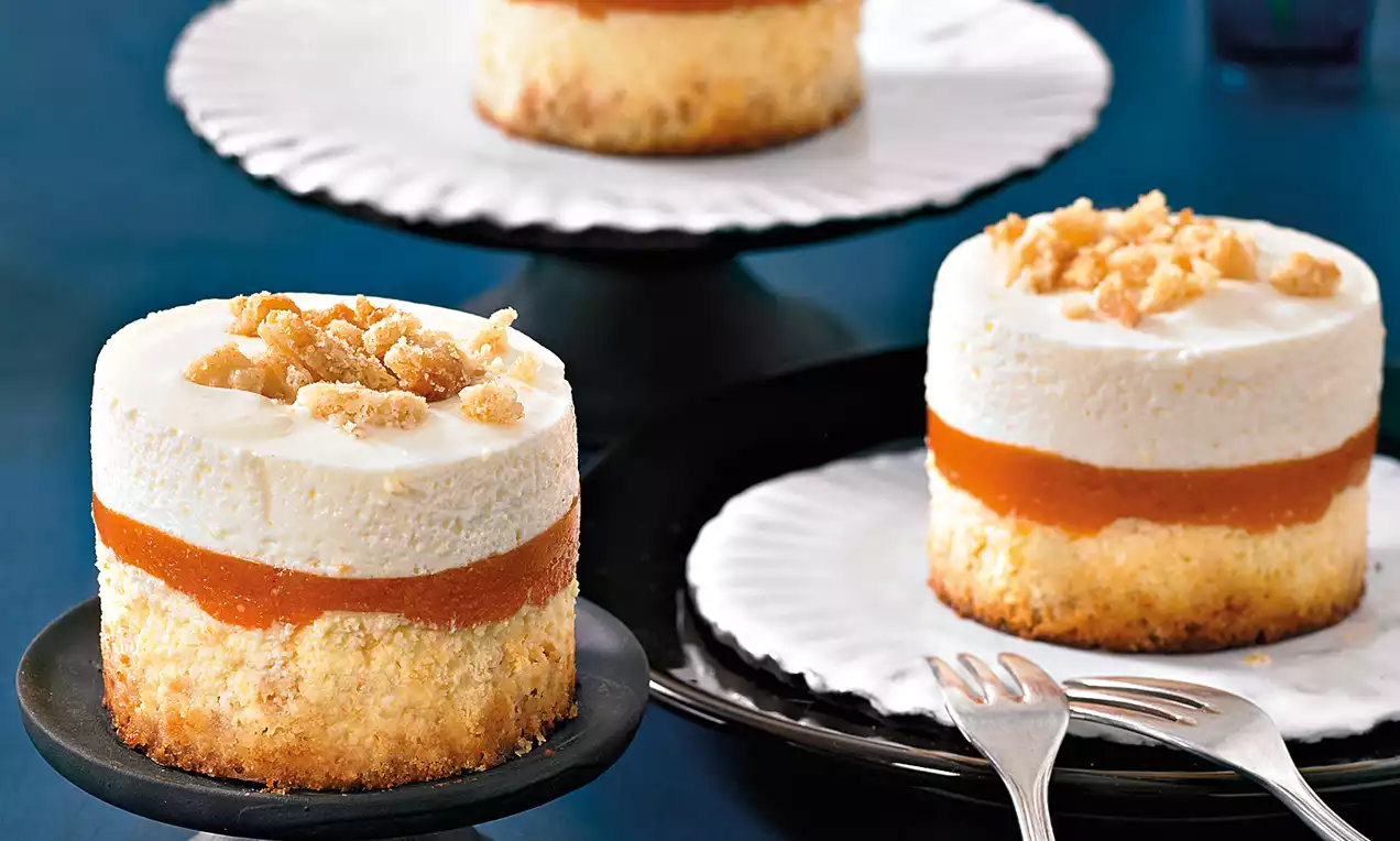 Mini-Aprikosen-Cheesecakes mit hellgoldenem Boden, leuchtend oranger Aprikosenschicht, weißer Creme und Krümeldekor auf schwarzen Tellern