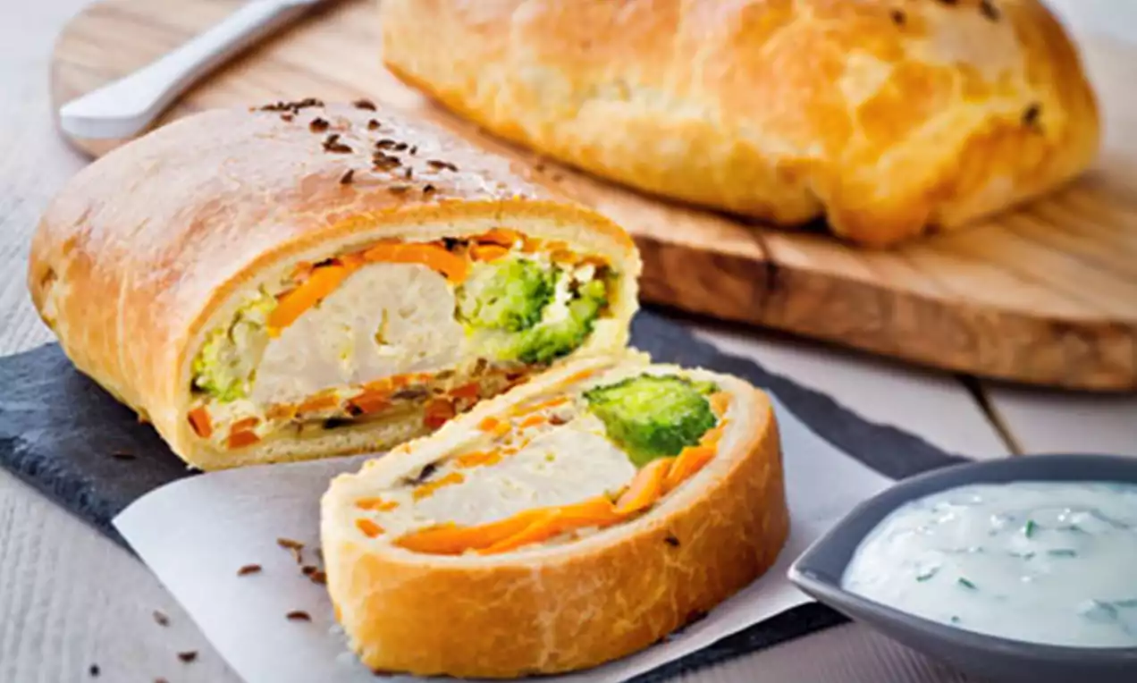 Złociste ciasto półkruche wypełnione warstwami marchewki, brokułów i innych warzyw, podane na ciemnej desce z sosem w miseczce