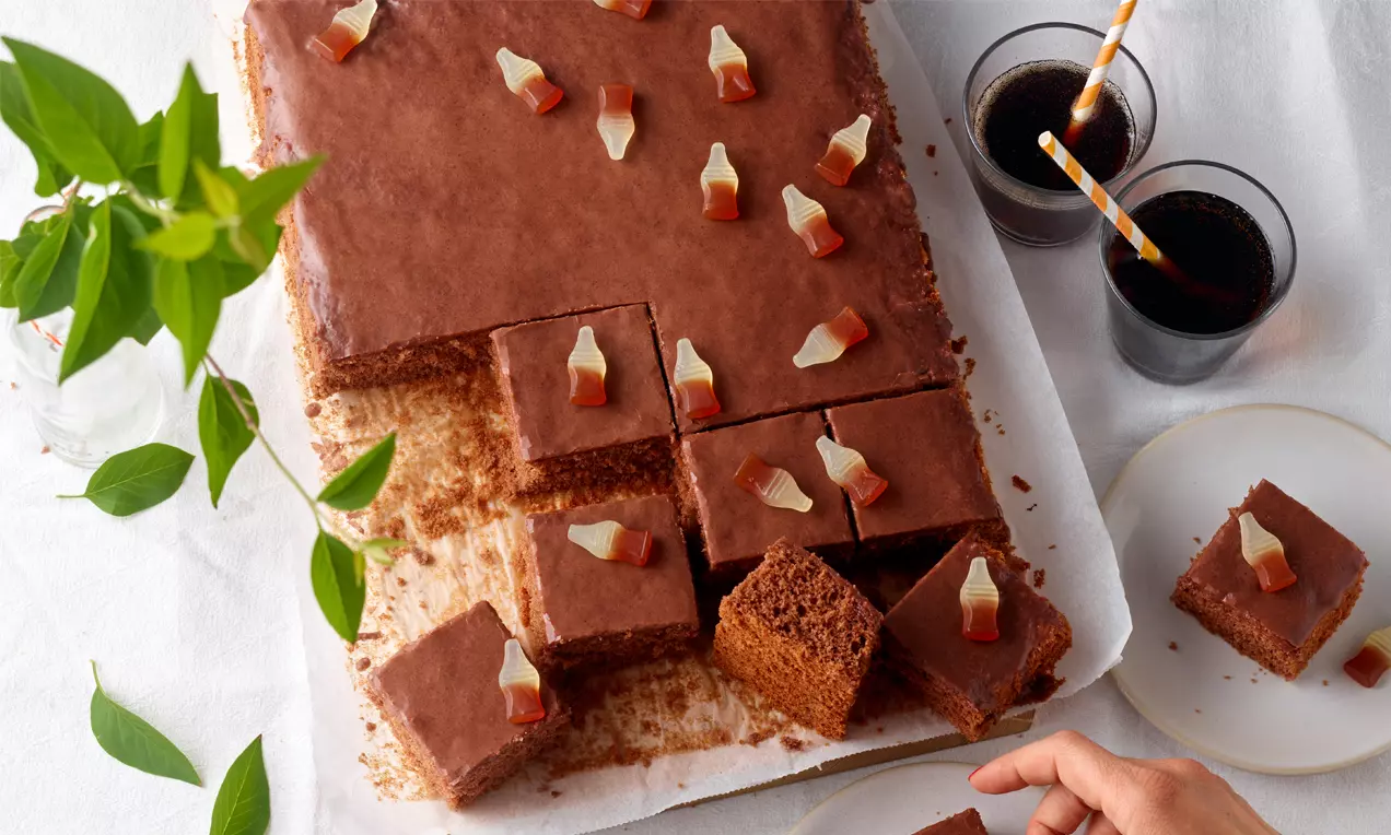 Rechteckiger Schokoladenkuchen mit glatter Glasur, garnierter Oberfläche aus kleinen Cola-Flaschen, geschnitten auf weißem Papier