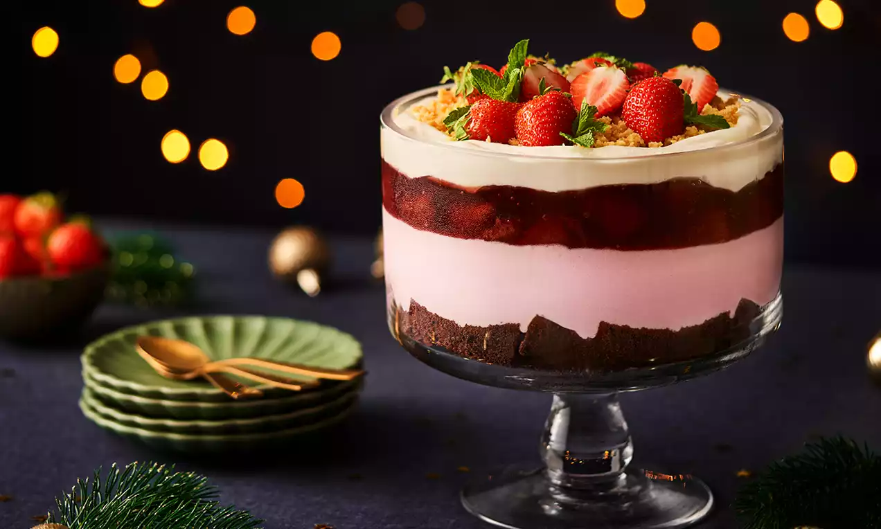 Glazen schaal met lagen chocoladecake, roze mousse, rode fruitsaus, vanillecrème, verse aardbeien en muntblaadjes, op een donkerblauwe feesttafel