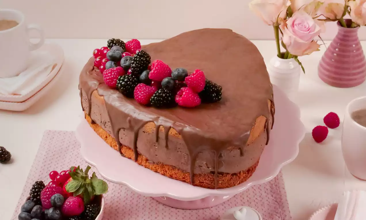 Schokoladen-Herzform mit glänzender Glasur, dekoriert mit frischen Himbeeren, Brombeeren und Blaubeeren auf rosa Tortenplatte