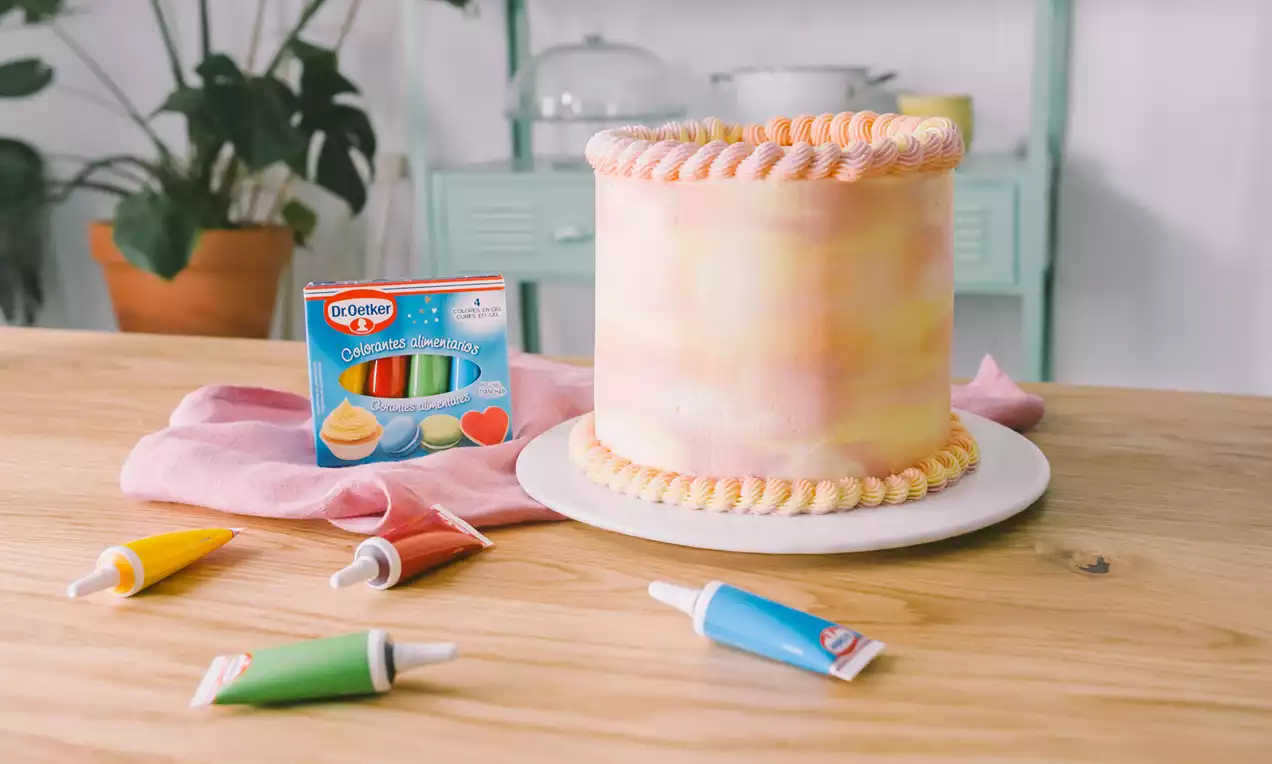 Pastel cilíndrico de tonos pastel con efecto acuarela, decorado con bordes de crema rizada, sobre un plato blanco en una mesa de madera