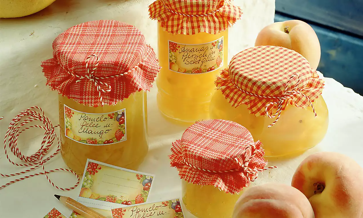 Gläser mit gelbem Pomelogelee, verziert mit rot-weiß karierten Stoffdeckeln und handgeschriebenen Etiketten, umgeben von frischen Pfirsichen