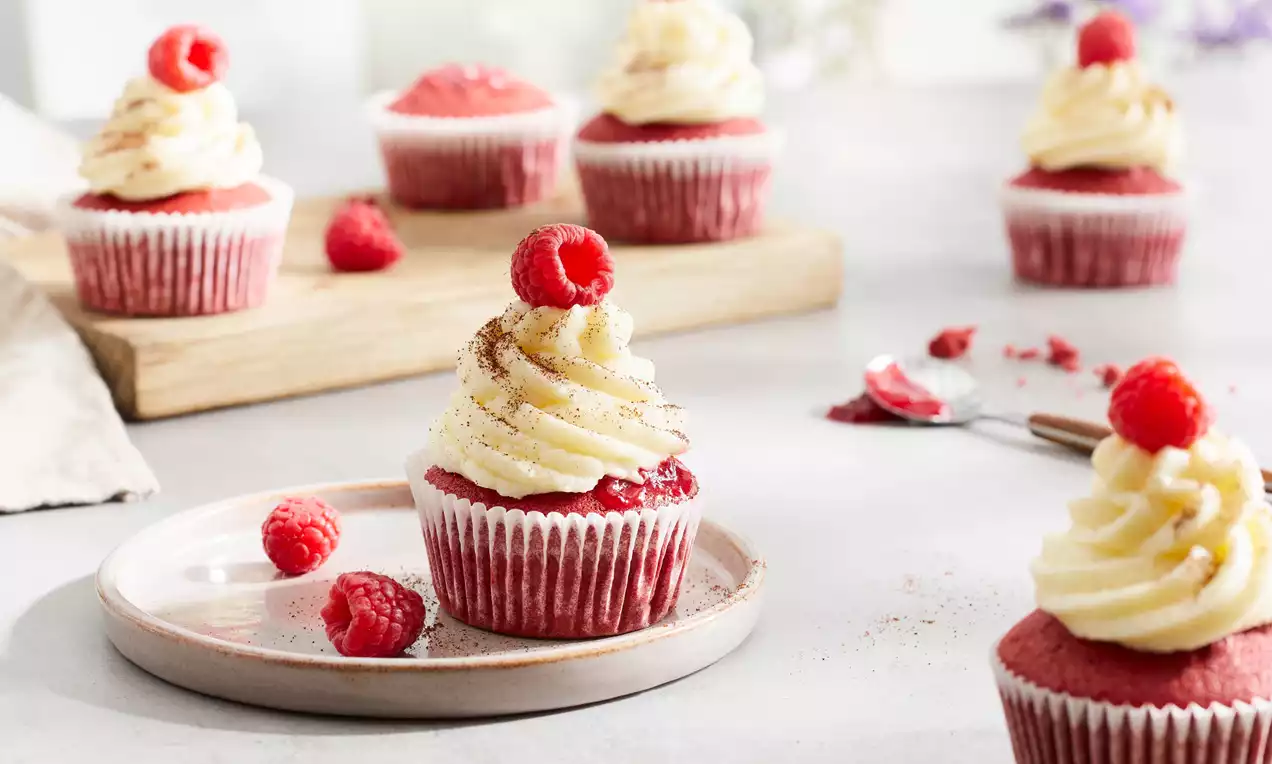 Frambozen cupcakes met rode cake, wit vanilleglazuur, verse frambozen en cacaopoeder, gepresenteerd op een licht bord en houten plank