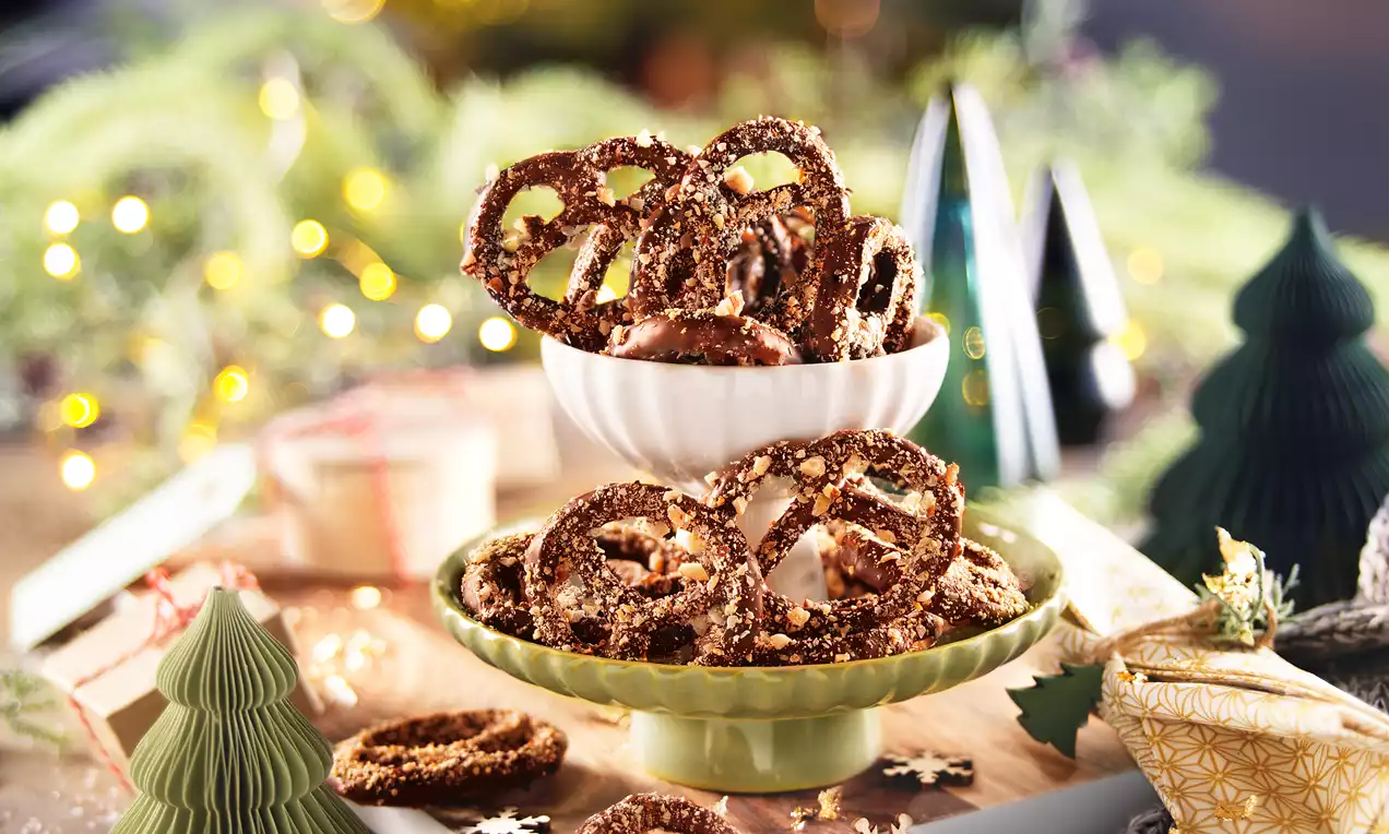 Maxi bretzels brillants au chocolat et éclats de noisettes dorées, disposés en pyramide dans des bols sur une table décorée de sapins