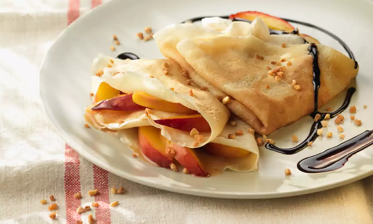 Crepe dorado relleno de rodajas de nectarina naranja y roja, doblado en un plato con hilo de chocolate y trozos de almendra sobre mantel claro