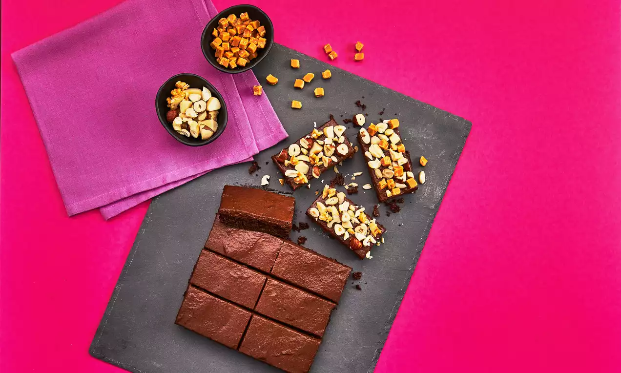 Dunkelbrauner Protein-Brownie in Rechtecken auf grauem Schiefer, mit Nüssen und orangefarbenen Stückchen garniert, neben Schalen und pinkem Tuch