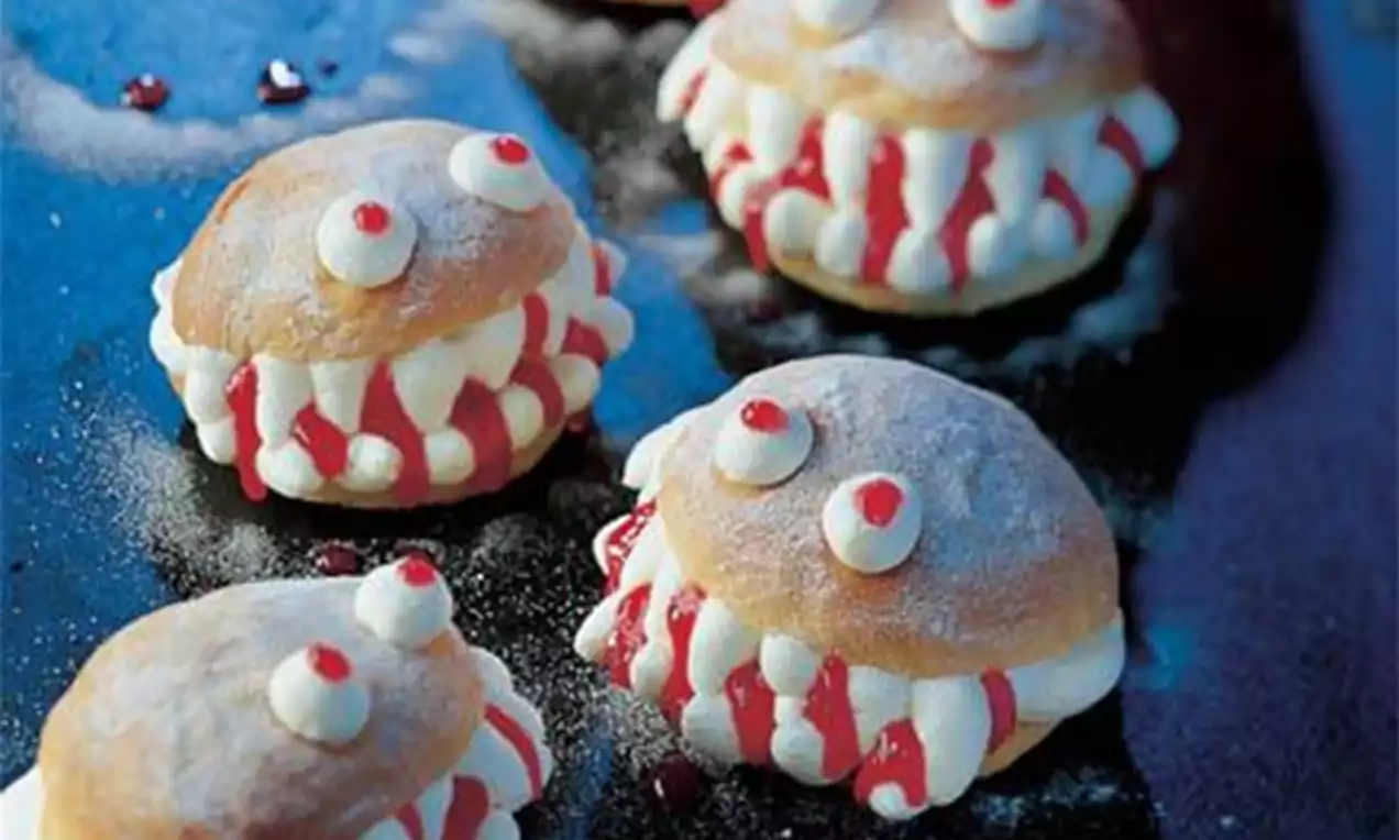 Petites brioches dorées rondes garnies de crème blanche et de coulis rouge, ornées de yeux en sucre sur une surface sombre