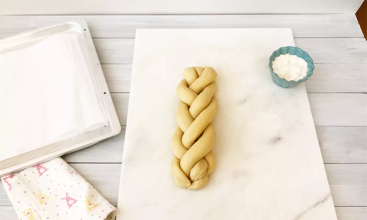 Светложълта плитка от тесто със стегнати усукани линии върху бял мраморен плот до синя купичка с брашно и дискретна шарена кърпа