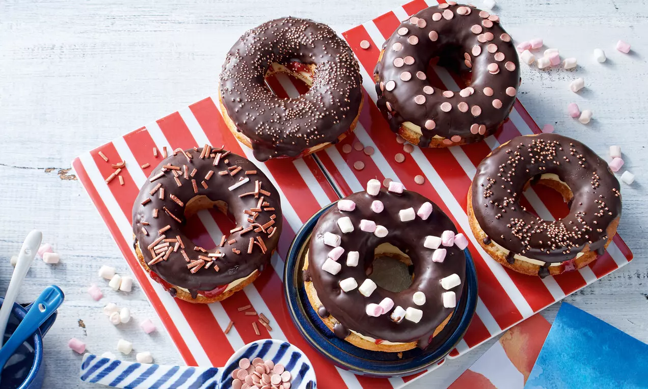 Кръгли донъти с тъмнокафява шоколадова глазура, поръсени с цветни пръчици и мини маршмелоу, подредени върху ярка червено-бяла основа