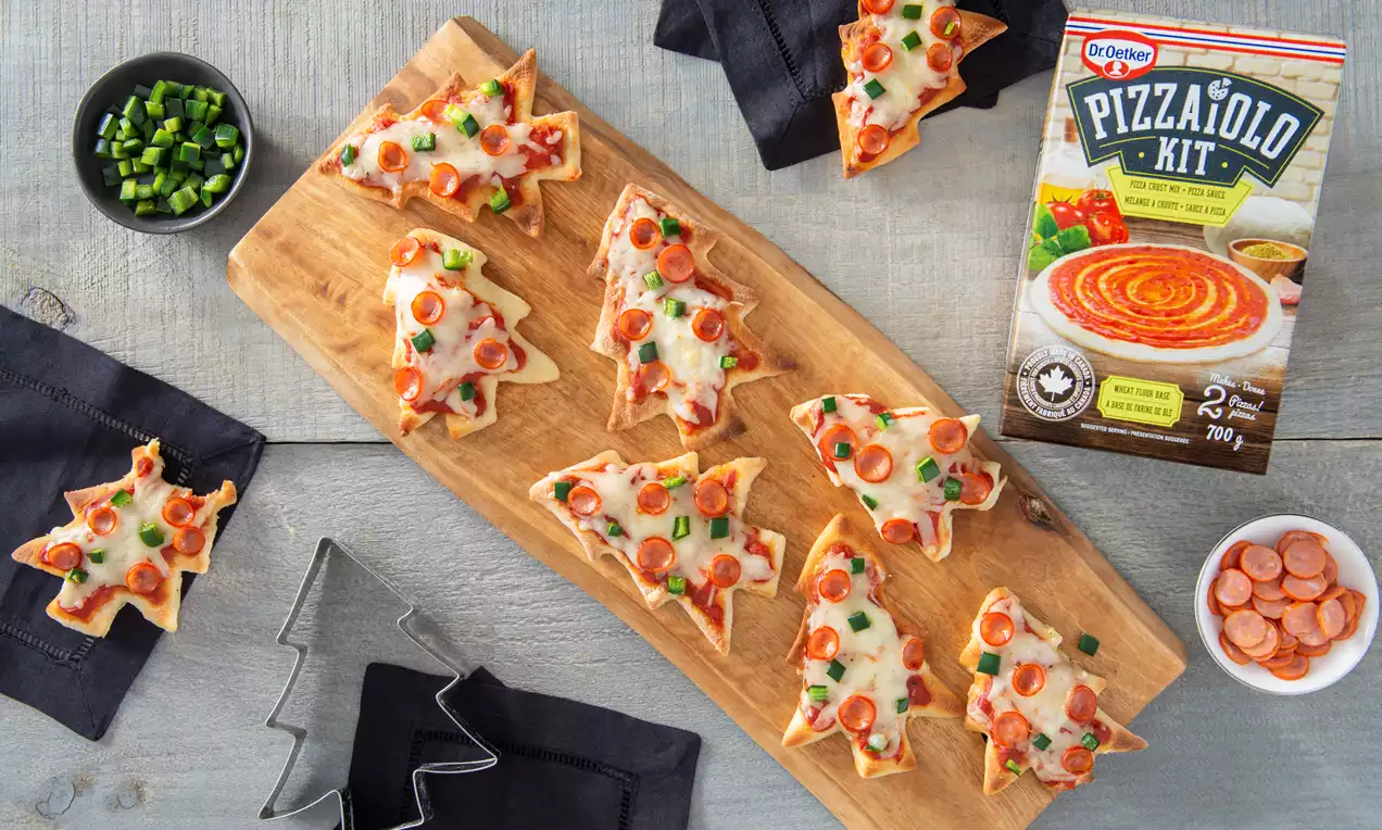 Mini tree-shaped pizzas with golden crust, melted cheese, red pepper slices, and green herbs on a wooden serving board, surrounded by toppings